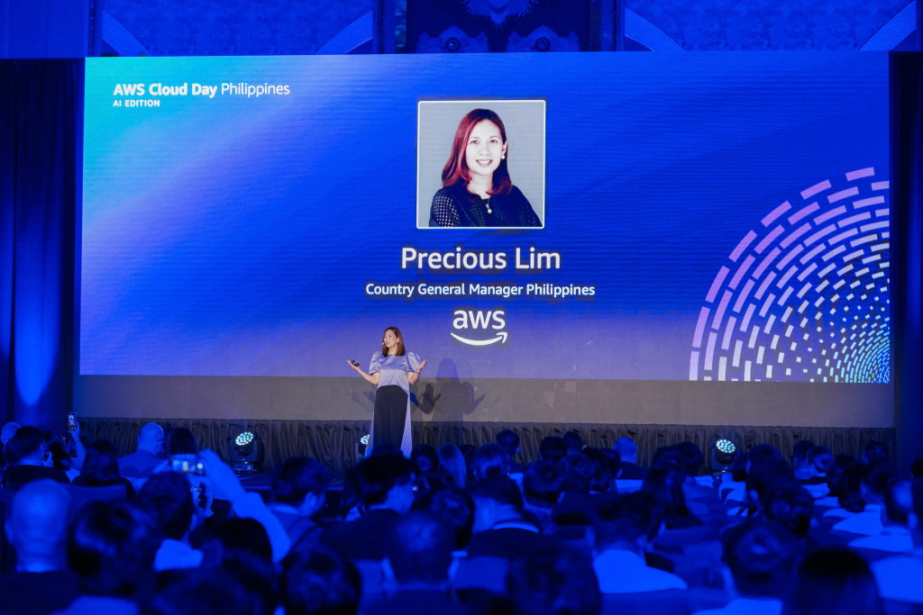 Speaker presenting on stage at AWS Cloud Day Philippines AI Edition, with a large screen showing Precious Lim, Country General Manager Philippines at AWS. The audience is seated in front, and the stage is lit with blue lighting and digital graphics.