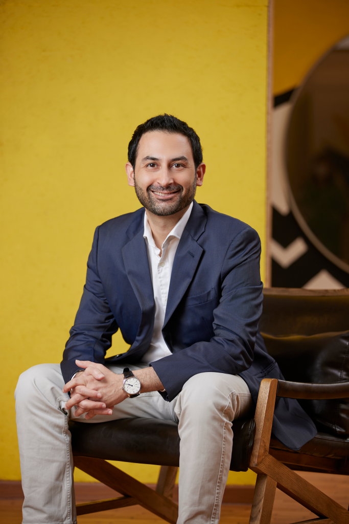 A man dressed in a suit is seated on a chair, positioned in front of a vibrant yellow wall.