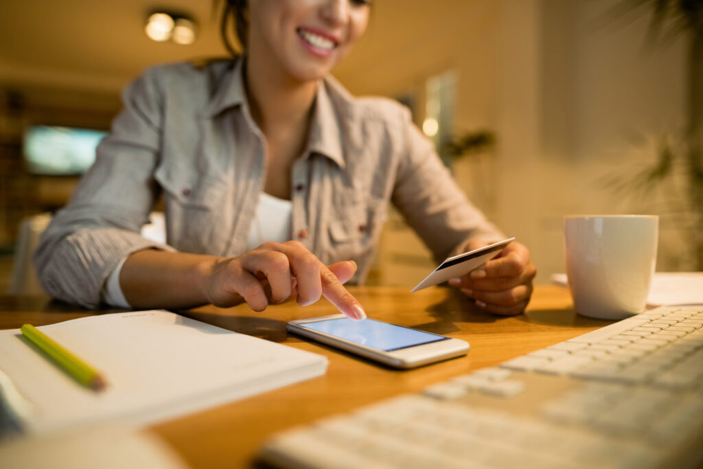 Woman using a smartphone for digital banking at home, holding a credit card with a notebook, keyboard, and coffee cup on the desk.