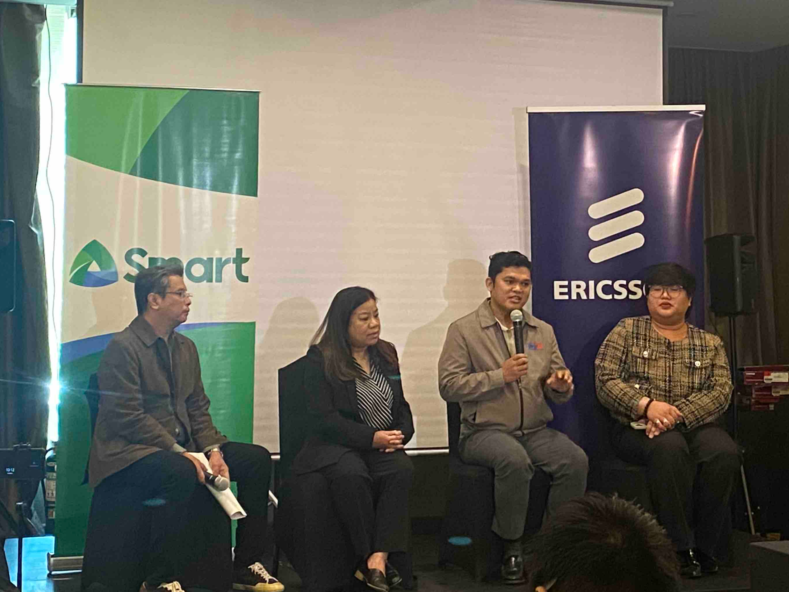 Roby Alampay, Vice President for Public Engagement and Corporate Communications at PLDT and Smart; Ellen Alarilla, Sustainability Manager for Ericsson SE Asia, Oceania and India; Dr. Jayboy Evano, Principal of the Sucat National High School; and Dr. Paul dela Vega,Senior Education Program Specialist in one frame.doing a Q&A session, with SMART and Ericssonn backdrop. Dr. Jayboy Evano holding a microphone while answering a question.