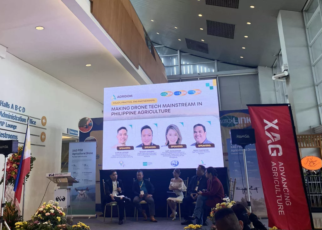 Panel discussion at an agriculture conference featuring a large digital screen displaying the topic “Making Drone Tech Mainstream in Philippine Agriculture” organized by AGRIDOM. Four speakers’ portraits and names are shown on the screen, while seated panelists discuss drone technology on stage. Banners for XAG agricultural drones and event sponsors are visible, along with signage for the administration office, restrooms, and VIP lounge.