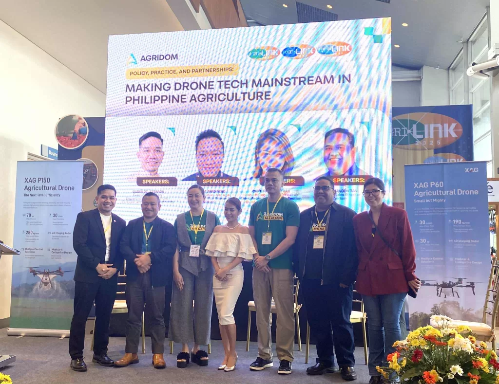 Group photo of speakers and participants at an agricultural technology event titled “Making Drone Tech Mainstream in Philippine Agriculture,” organized by AGRIDOM. The large digital screen behind them displays speaker photos and event logos, while banners for XAG P150 and XAG P60 agricultural drones stand on either side. The group poses indoors with floral decorations in front.