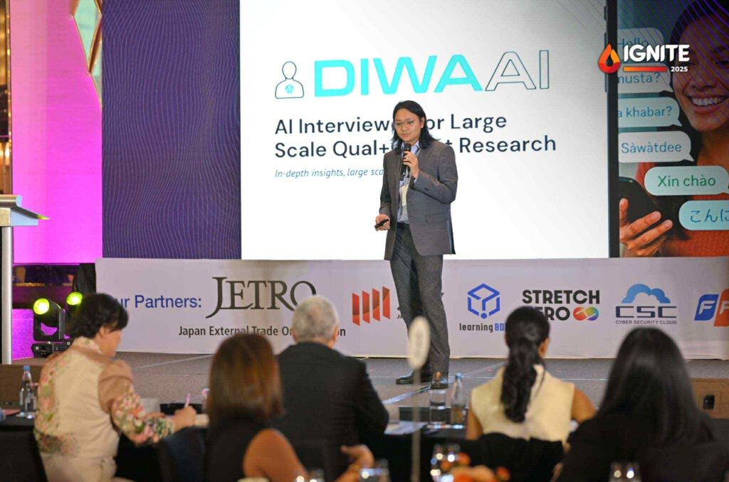 A speaker presents on stage at the IGNITE 2025 event with a large screen displaying “DIWA AI – AI Interviews for Large Scale Qual+Quant Research.” The audience is seated at tables, and partner logos including JETRO, Stretch Pro, and CSC are visible on the stage backdrop.