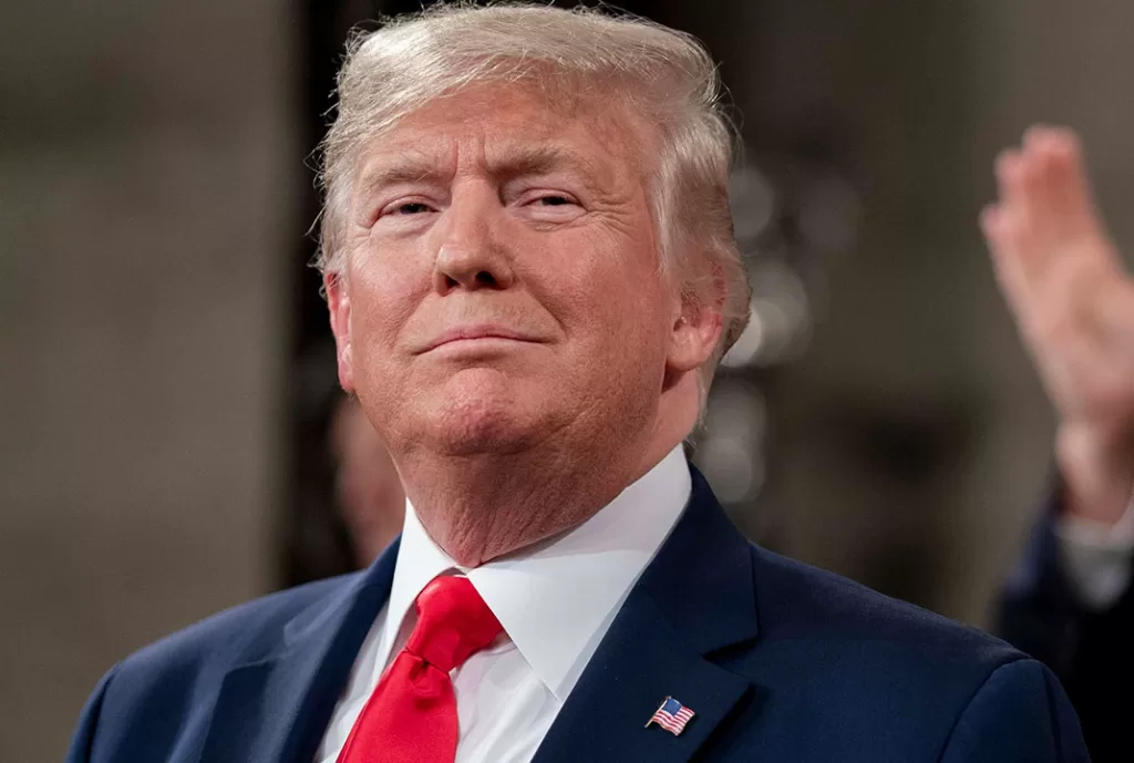 A serious-looking President Donald Trump speaks at a press conference, standing behind a podium with U.S. flags in the background. His expression is firm but guarded, as he addresses questions about the potential economic impact of his new tariff policies. Reporters’ microphones and cameras are visible in the foreground. The atmosphere feels tense, reflecting market uncertainty and public concern over whether the tariffs could trigger a recession.