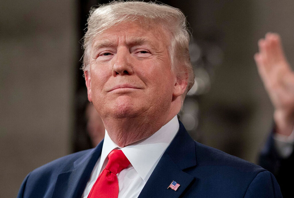 A serious-looking President Donald Trump speaks at a press conference, standing behind a podium with U.S. flags in the background. His expression is firm but guarded, as he addresses questions about the potential economic impact of his new tariff policies. Reporters’ microphones and cameras are visible in the foreground. The atmosphere feels tense, reflecting market uncertainty and public concern over whether the tariffs could trigger a recession.