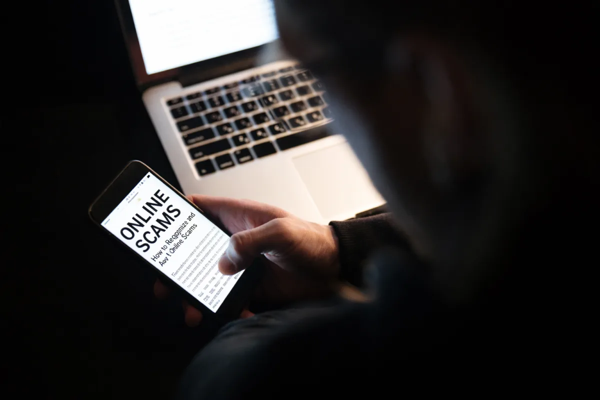 Online scams are becoming rampant as shown in this image of a man using laptop computer and phone; written on the screen are the words "online scams"
