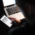 Online scams are becoming rampant as shown in this image of a man using laptop computer and phone; written on the screen are the words "online scams"