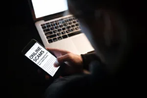 Online scams are becoming rampant as shown in this image of a man using laptop computer and phone; written on the screen are the words "online scams"