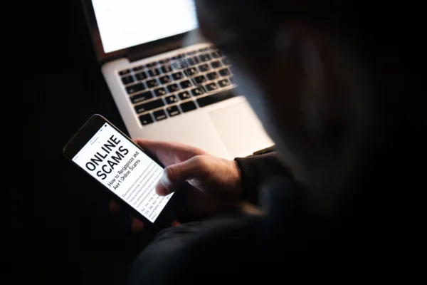 Online scams are becoming rampant as shown in this image of a man using laptop computer and phone; written on the screen are the words "online scams"